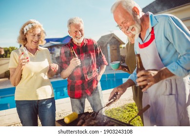 Group Of Senior Friends Having A Backyard Barbecue Party By The Swimming Pool, Gathered Around The Grill, Grilling Meat And Having Fun