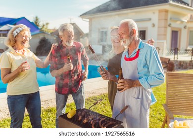 Group Of Senior Friends Having A Backyard Barbecue Party By The Swimming Pool, Gathered Around The Grill, Grilling Meat And Having Fun