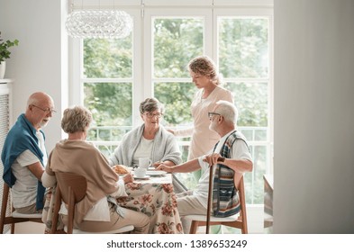 Group Of Senior Friends With Helpful Carer Sitting Together At The Table At Nursing Home Dining Room