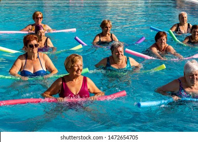Group Of Senior Ladies Doing Exercise In Outdoor Swimming Pool. Aqua Gym Class In Action With Foam Noodles.
