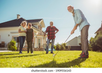 Group Of Senior Neighbors Gathered Playing Mini Golf At The Backyard Lawn, Having Fun Spending Sunny Summer Day Outdoors