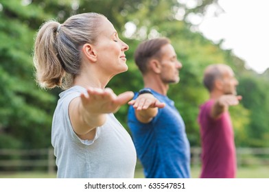 Group of senior people with closed eyes stretching arms at park. Happy mature people doing yoga exercise outdoor on a bright morning. Yoga class with woman and men doing breath exercise.