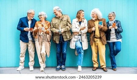 Group of senior people using technology devices together standing on a blue wall - Happy older friends having fun watching funny video on smartphone - Tech and modern elderly concept