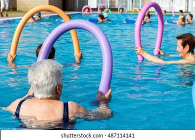 Group Of Senior Women Doing Rehabilitation Exercise With Soft Foam Noodles In Outdoor Swimming Pool.