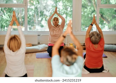 A group of senior women engage in various yoga exercises, including neck, back, and leg stretches, under the guidance of a trainer in a sunlit space, promoting well-being and harmony - Powered by Shutterstock