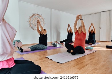 A group of senior women engage in various yoga exercises, including neck, back, and leg stretches, under the guidance of a trainer in a sunlit space, promoting well-being and harmony - Powered by Shutterstock