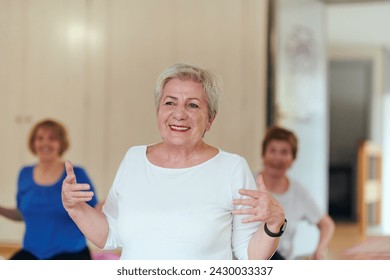 A group of senior women engage in various yoga exercises, including neck, back, and leg stretches, under the guidance of a trainer in a sunlit space, promoting well-being and harmony - Powered by Shutterstock