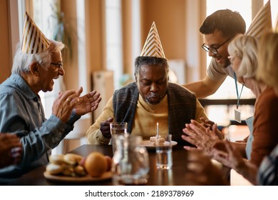 Group Of Seniors Celebrating African American Man's Birthday While He Is Blowing A Candle In Nursing Home.