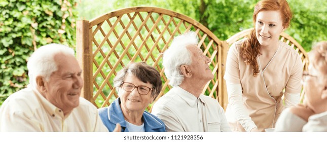 Group Of Seniors Enjoying Their Time Together Outside In A Garden Of An Assisted Living Home. A Young Volunteer Helping. Panorama.