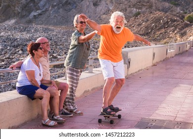 Group Of Seniors And Mature People At The Beach Have Fun Looking At Old Man Riding A Skateboard And Laughing With Scare Face - Woman Touching The Man