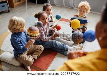Group of serious intercultural learners of primary school holding models of solar system planets while sitting on the floor in front of teacher
