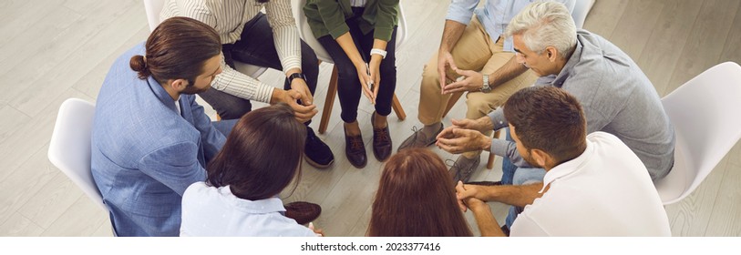 Group Of Serious People Sitting In A Circle During A Therapy Session. Young And Mature Men And Women Meeting For A Conversation With A Psychologist. Overhead View, High Angle Shot, Banner Background