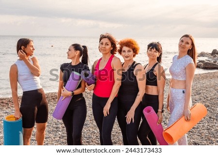 Group of six Caucasian adult women posing holds sports mats on pebble wild beach. Sea and wild coast in background. Fitness lifestyle outdoor training.
