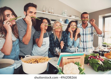 Group Of Six Diverse Silly Adults Sniffing Asparagus Stalks In Kitchen. Bowl Of Pasta And Vegetables Are On The Table.