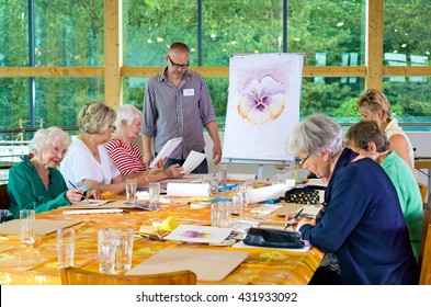 Group of six female senior citizens at long table in painting class with male teacher in glasses standing near front - Powered by Shutterstock