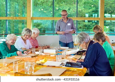 Group Of Six Female Senior Citizens At Long Table In Art Class With Male Teacher In Glasses Standing Near Front Beside Large Windows Facing Green Trees