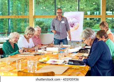 Group of six female senior citizens at long table in painting class with male teacher in glasses standing near front - Powered by Shutterstock