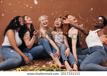 Group of six laughing women of different ages sitting under falling flower petals. Multi-ethnic smiling females having fun in studio while sitting on brown background.