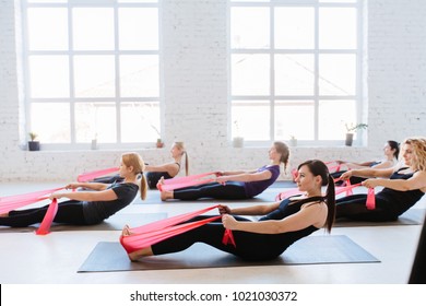 Group of six women are doing stretching exercise with red resistance bands in white studio interior. Teamwork, good mood and healthy lifestyle concept. - Shutterstock ID 1021030372
