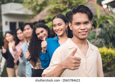 A group of six young and dynamic friends from late teens to early 20s, in a single file giving a thumbs up. Outdoor scene.