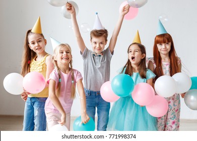 Group of small children have birthday party, wear festive hats, hold balloons, have joy together, enjoy playing games. Small adorable girl celebrates her birthday, invites friends, have happy looks