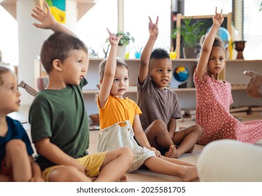 Group of small nursery school children sitting on floor indoors in classroom, raising hands. - Shutterstock ID 2025581705