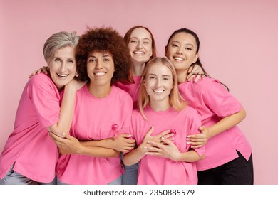 Group of smiling confident multiracial women wearing t shirts with pink ribbon looking at camera isolated on pink background. Health care, support, prevention. Breast cancer awareness month concept