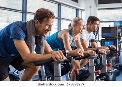 Group of smiling friends at gym exercising on stationary bike. Happy cheerful athletes training on exercise bike. Young men and woman working out at a class in the gym.