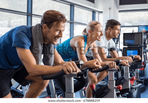 Group of smiling\
friends at gym exercising on stationary bike. Happy cheerful\
athletes training on exercise bike. Young men and woman working out\
at a class in the gym.