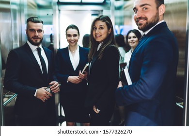 Group Of Smiling Successful People In Suits Carrying Documents While Standing Together And Looking At Camera In Elevator Cabin At Urban Office Building