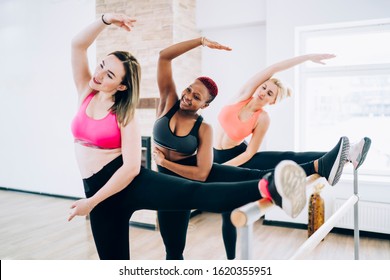 Group of smiling toned multiethnic sportswomen standing with one leg on barre and reaching over leg with opposite arm in bright fitness room - Powered by Shutterstock