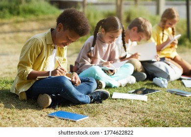 Group of smiling young children studying outdoors sitting in row on green grass and writing in textbooks, focus on African-American boy in foreground, copy space - Powered by Shutterstock