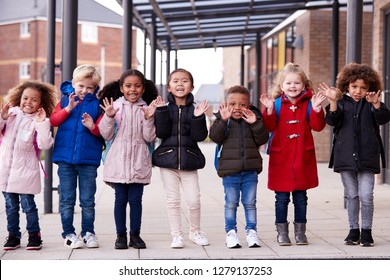 A Group Of Smiling Young Multi-ethnic School Kids Wearing Coats And Carrying Schoolbags Standing In A Row In Walkway Outside Their Infant School Waving To Camera, Full Length, Front View