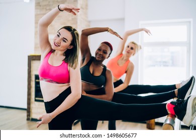 Group of sportive young female athletes wearing sports bra and leggings smiling and stretching using ballet barre during ballet class in studio