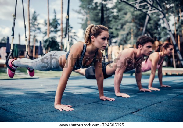 Group of sports people are\
training outdoors with TRX. Total Body Resistance Exercises. Two\
attractive young women and handsome man are working out in\
gym.
