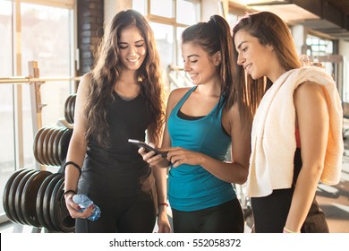 Group of sporty female friends using app on mobile phone, looking at screen of smart phone, texting, watching photos while resting after practice, on break between exercises in gym. - Powered by Shutterstock