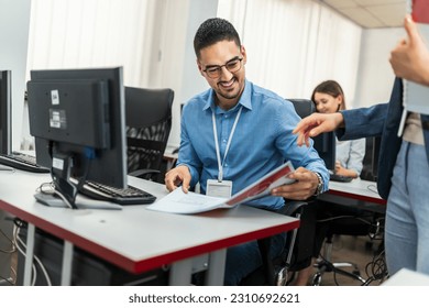A group of students each sit in front of a desktop in a computer lab. They are each focused on their screens as their female teacher gives them scripts for class. 