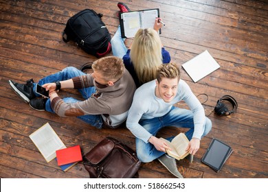 Group of students reading books, writing in notebooks and using a smartphone leaning on each other on wooden floor. 