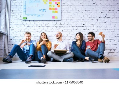 Group Of Students Sitting On Floor In Campus And Eating Pizza Together