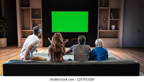 A Group Of Students Is Watching A TV And Celebrating Some Joyful Sports Moment, Sitting On The Couch In The Living Room. The Living Room Is Made In 3D. TV Is Green Screen For Further Editing.