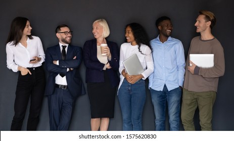 Group Of Successful Happy Diverse Young And Older Business People Staff With Gadgets Standing In Line Near Black Wall, Communicating, Talking, Having Fun Together, Human Resource Team Concept.