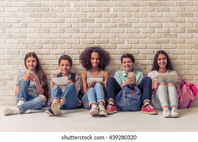 Un grupo de adolescentes está usando aparatos, mirando la cámara y sonriendo, sentados contra una pared de ladrillo blanco
