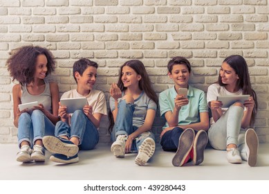 Group of teenage boys and girls is using gadgets, talking and smiling, sitting against white brick wall