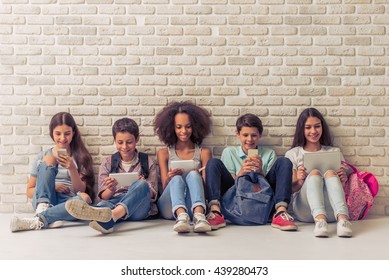 Group of teenage boys and girls is using gadgets and smiling, sitting against white brick wall
