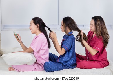 Group Of Teenage Girls Having Sleepover Party While Tying Hair To Each Other And Sitting On The Bed At Home