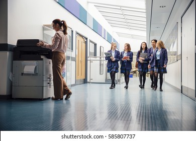 Group Of Teenage Girls Are Walking Down The School Hall With Books And Laptops In Their Arms. They Are Talking And Laughing As They Walk And There Is A Female Teacher Using The Printer.