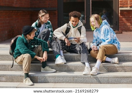 Group of teenage students sitting outside school buildings and watching funny video on smartphone