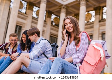 A group of teenage tourists are resting sitting on the steps into the museum. Guys are watching something on the phone while the girl is happily chatting on the phone. - Powered by Shutterstock