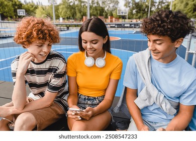 Group of teenagers high school pupils classmates college students using gadgets mobile app watching video holding smart phone play video game in sport basketball court yard sitting outside - Powered by Shutterstock