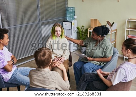 Group of teenagers sitting on chairs and talking their problems to psychologist who listening to them and giving advice during psychology therapy group